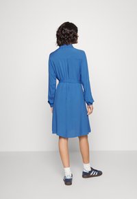 Abito blu a maniche lunghe con vita cintata, tessuto morbido e orlo leggermente svasato. Abbinato a sneakers scure e calzini bianchi.