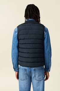 Chaleco de plumas negro con costuras horizontales, llevado sobre una camisa de mezclilla. La parte de atrás presenta un cuello alto y una textura mate suave.