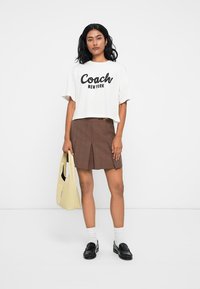 Camiseta blanca corta con el logo "Coach New York", falda de cuadros marrón con pliegues, mocasines negros, calcetas blancas y bolso tote de color amarillo pálido.