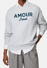 Grijze sweatshirt met de tekst "AMOUR Parijs" in donkerblauwe borduursel. Draagt over een wit overhemd met een kraag en strepen op grijze broek.