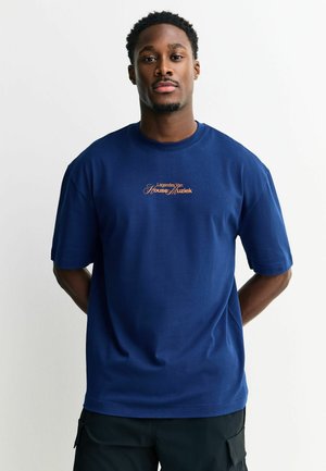 Mees seisab käed selja taga, kandes sinist T-särki, millel on rinnal tekst "Legendes Van House Muziek".