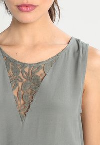 Blouse grise sans manches avec un décolleté en V orné d'un détail en dentelle florale, texture douce et design épuré sans ornements.