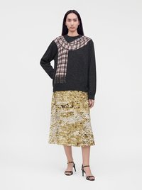 Femme debout portant un pull sombre avec un motif d’écharpe imprimé et une jupe midi à sequins dorés avec des sandales noires à talons hauts.
