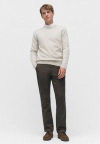 Lys beige sweater med ribbede manchetter og kant, sammen med mørkebrune bukser og brune sko, stående mod en grå baggrund.