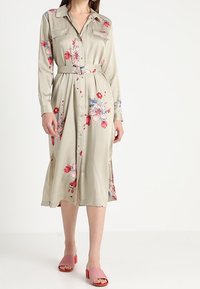 Vestito a maniche lunghe, con bottoni, di colore verde chiaro con stampa floreale. Caratterizzato da una vita con cintura, tessuto morbido e spacchi laterali. Indossato con sandali con tacco a blocco rosa.