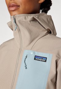 Bužinio spalvos zip-up švarkas su aukštu apykakle, turintis mėlyną krūtinės kišenę ir logotipo pleistrą. Lygus audinys su zigzago raštu.