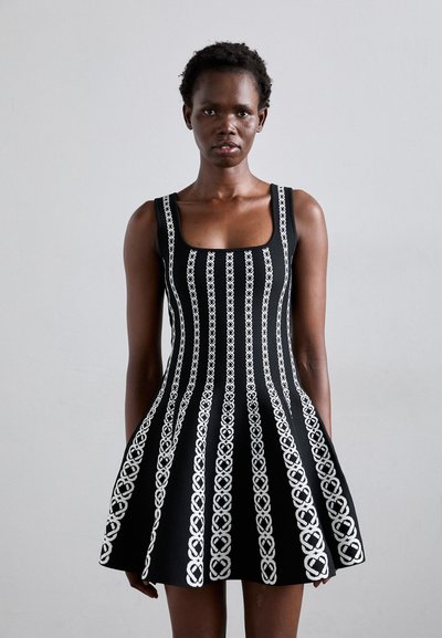 Robe noire sans manches avec un décolleté carré, ornée de motifs en forme de cœurs blancs en rayures verticales, évasée au niveau de la jupe.