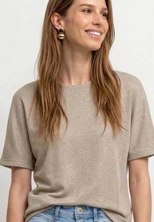 Jeune femme aux longs cheveux bruns portant des boucles d'oreilles rondes dorées et un haut beige scintillant à manches courtes rentré dans un jean bleu, souriante et regardant vers la droite.
