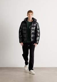 Zwarte pufferjack met een glanzende afwerking, gequilt ontwerp, met capuchon, gedragen over een groene T-shirt en zwarte broek; model draagt witte sneakers.
