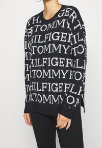 Marineblauer Strickpullover mit weißem "TOMMY HILFIGER"-Muster, geripptem Ausschnitt und Bündchen, seitlichen Schlitzen und kleinem Logo-Akzent am Ärmel.