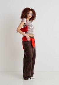 Γυναίκα με σγουρά μαλλιά που φορά γυαλιά, ριγέ κροπ τοπ, φαρδιά καφέ παντελόνια, λευκά παπούτσια, κόκκινο πουλόβερ δεμένο γύρω από τη μέση και κρατά κόκκινη τσάντα.