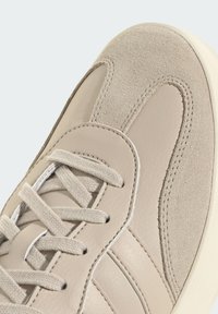 Beige sneaker close-up die suède en leertexturen toont, met gestikte overlappen en platte geweven veters op een lichte zool.