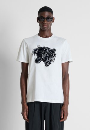 Man draagt een zwarte zonnebril, wit t-shirt met print van een brullende zwarte panter en losse zwarte broek, staand tegen een effen achtergrond.