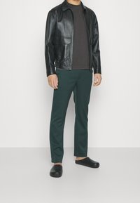 Chaqueta de cuero negra con dos bolsillos frontales, camiseta marrón oscuro, pantalones verdes ajustados y zapatos negros sin cordones. Texturas suaves, diseño elegante.