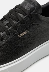 Zapatilla de cuero negra con superficie texturizada, cordones planos negros y suela de goma blanca con un acento metálico.