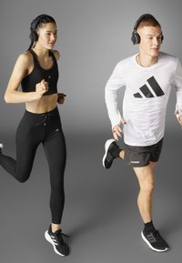 Kaksi henkilöä, jotka käyttävät kuulokkeita, juoksevat urheilullisissa Adidas-vaatteissa; toinen mustissa leggings-housuissa ja urheiluliivissä, toinen mustissa shortseissa ja valkoisessa pitkähihaisessa paidassa.