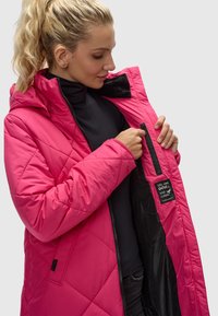 Giacca isolante rosa con un motivo a rombi, cappuccio e tasca interna con zip. L'interno presenta un rivestimento nero e un'etichetta con il logo.