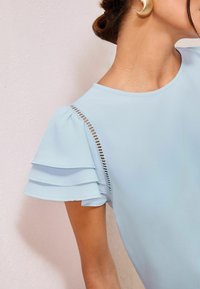 Blusa azzurra chiara con maniche corte a volant, caratterizzata da un dettaglio a taglio lungo la cucitura della spalla. Tessuto morbido con scollatura rotonda.