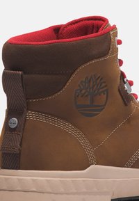 Botte montante en cuir marron avec un col en textile rouge, surpiqûres blanches, logo noir et œillets en métal pour les lacets, dotée d'une semelle texturée.