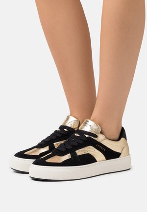 Pieds chaussés de baskets basses noires et dorées avec semelles blanches et lacets noirs, posés sur un fond clair uni.