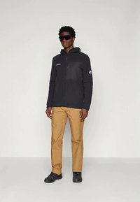 Mees, kes kannab musta tõmblukuga kapuutsi, tumedaid pükse ja musti kingi, seisab tavalise heleda tausta vastu, suurte tumedate prillide peal.