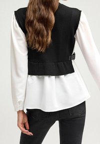 Gilet court noir avec une texture côtelée, associé à une chemise blanche. Le gilet a un ourlet peplum et un détail de boucle ajustable au dos.