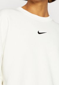 Vit sweatshirt i mjukt tyg, med rund halsringning och en liten svart Nike-logga på bröstet. Slät textur, avslappnad passform.