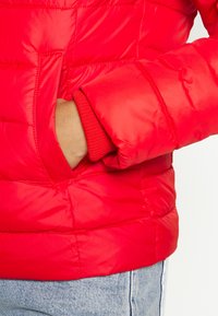 Red puffer jacket shows quilted design with stitched sections and elastic cuffs. Pocket visible on the left side, paired with denim jeans.