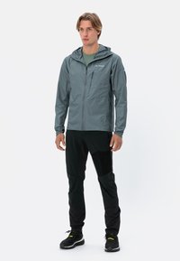 Giacca leggera e impermeabile di colore grigio, con zip frontale, cappuccio regolabile e tasca sul petto, indossata sopra pantaloni neri e scarpe da ginnastica nere.