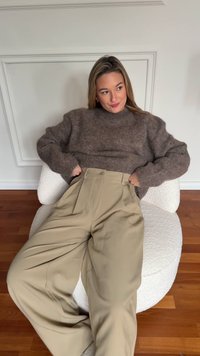 Pruun tekstuuriga kampsun kõrge kaelusega, kombineeritud kõrge vöökohaga beežide volditud pükstega, istumas valgel tekstuuriga toolil.