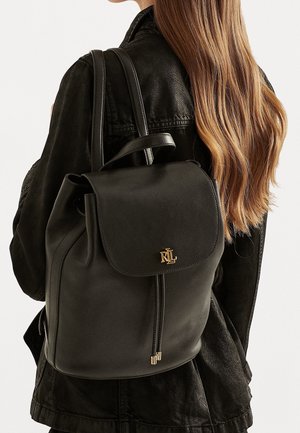 Svart läderryggsäck med klaffstängning, guldlogotyp som accent och justerbara remmar. Slät textur och rund form med en stilren design.
