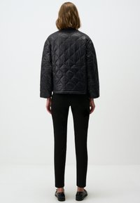 Veste noire matelassée avec un col rond et des manches longues, associée à un pantalon noir slim et des chaussures foncées, vue de l'arrière.