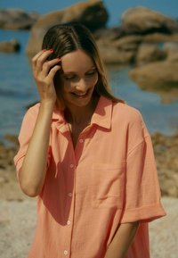 Leichtes, korallenfarbenes Button-up-Hemd mit einer Tasche, umgeschlagenen kurzen Ärmeln und lässiger Passform, vor einem felsigen Strandhintergrund.