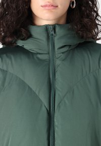 Grønn polstret jakke med høy krage, som har en full-lengde glidelås og en teksturert overflate. Stoffet har et glatt, vannavvisende utseende.