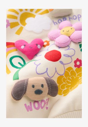 Flødefarvet stof med farverige broderede motiver. Bemærkelsesværdige detaljer omfatter en pink blomst, et hjerte, en hund med ører og solmotiver.