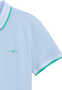 Hellblaues Baumwoll-Polohemd mit einem grün-weißen Kragen. Besitzt ein kleines, grünes Logo mit "1962" im Bereich der linken Brust.