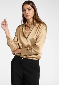 Frau trägt ein glänzendes goldenes Satin-Hemd mit Knöpfen und schwarze Hosen, posiert mit einem Arm verschränkt und dem anderen nahe ihrem Gesicht erhoben.