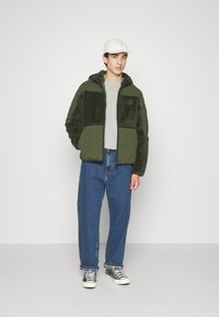 Veste en polaire verte avec des panneaux matelassés, t-shirt gris, jeans bleus amples et baskets grises. Casquette blanche avec un logo floral.