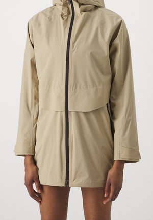 Beige regenjas met capuchon, zwarte rits aan de voorkant en lange mouwen, gedragen door een persoon met zichtbare handen en bovenbenen tegen een witte achtergrond.