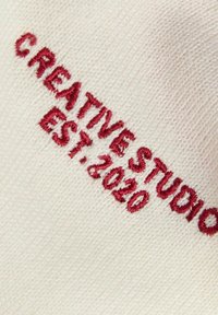Rød broderet tekst "CREATIVE STUDIO EST. 2020" på hvidt tekstureret stof.
