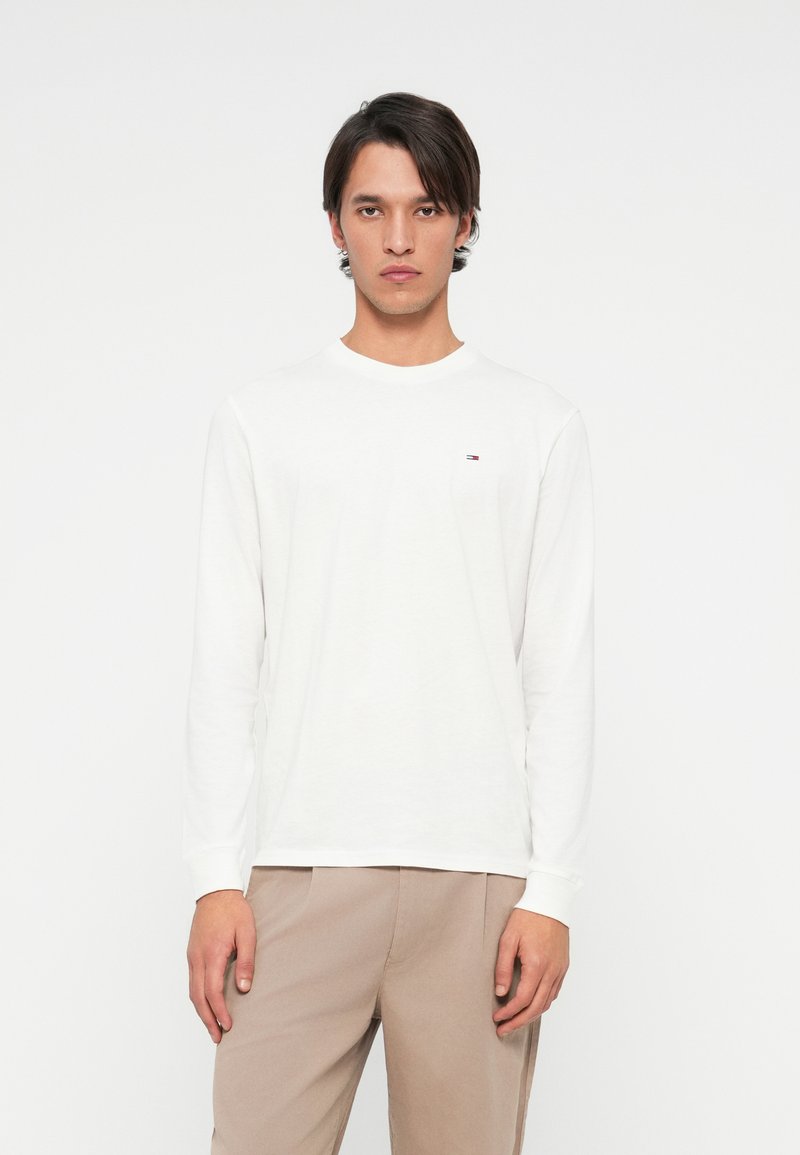 Joven con cabello oscuro y lacio, vistiendo una camisa blanca de manga larga y pantalones beige, de pie frente a un fondo blanco liso.