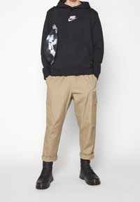 Svart hoodie med en rosa Nike-logga, beige cargo-byxor och svarta snörskor. Har också en sling-väska i kamouflage med Nike-branding.