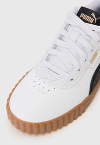 Baskets blanches avec une tige en cuir texturé, accents noirs et dorés, semelle en caoutchouc arrondie et lacets plats avec des détails perforés.
