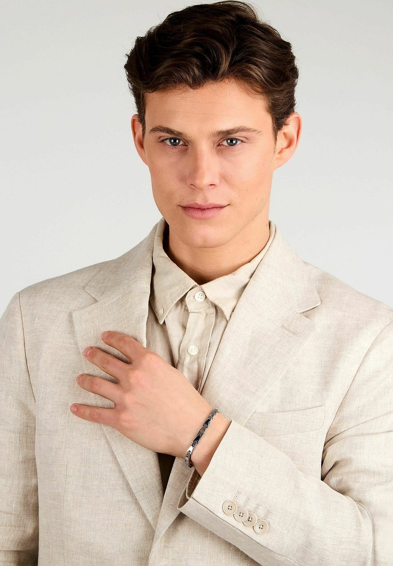 Bracciale d'argento su una mano, indossato con un completo in lino di colore beige chiaro e una camicia coordinata. Presenta motivi sottili e texture lisce.