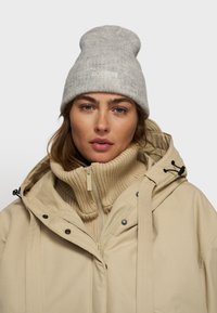 Lys grå uldbeanie med ribbet tekstur og logo-detalje, båret med en cremefarvet højhalset sweater og en beige vandtæt jakke.