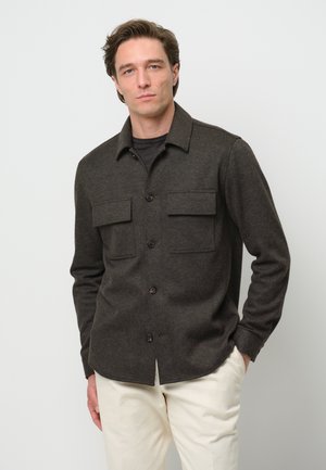 Chaqueta de camisa de lana gris oscuro con botones en el frente, dos bolsillos en el pecho y mangas largas, combinada con pantalones beige claros.