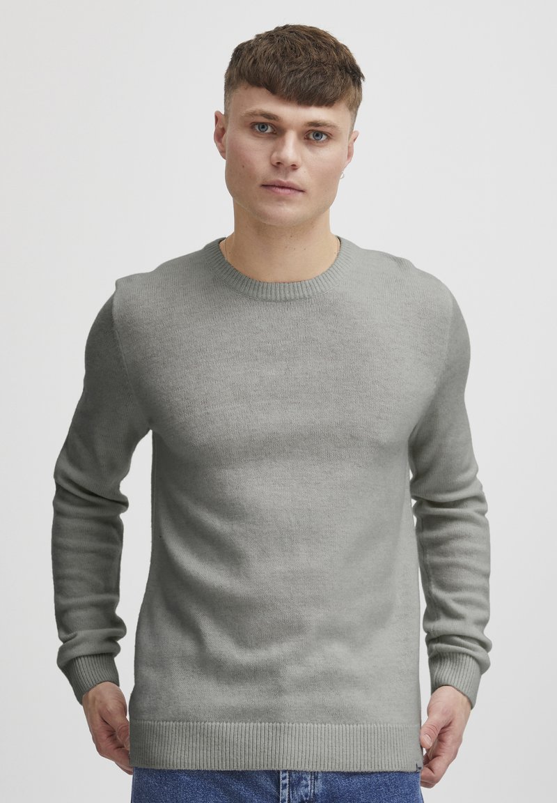 Pull en tricot gris clair avec un col rond, des manches longues et des bords côtelés aux poignets et à l'ourlet. Texture douce, coupe ajustée, détails minimes.