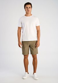 Biała koszulka z okrągłym dekoltem w zestawieniu z oliwkowozielonymi krótkimi spodniami. Modelka ma na sobie białe tenisówki. Prosty fason, wygodne dopasowanie, casualowy styl.