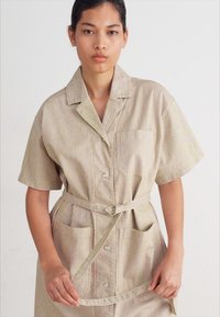 Stripet skjortedress i beige og krem, med korte ermer, krage, frontknapper, og en knyttet midje med to frontlommer.