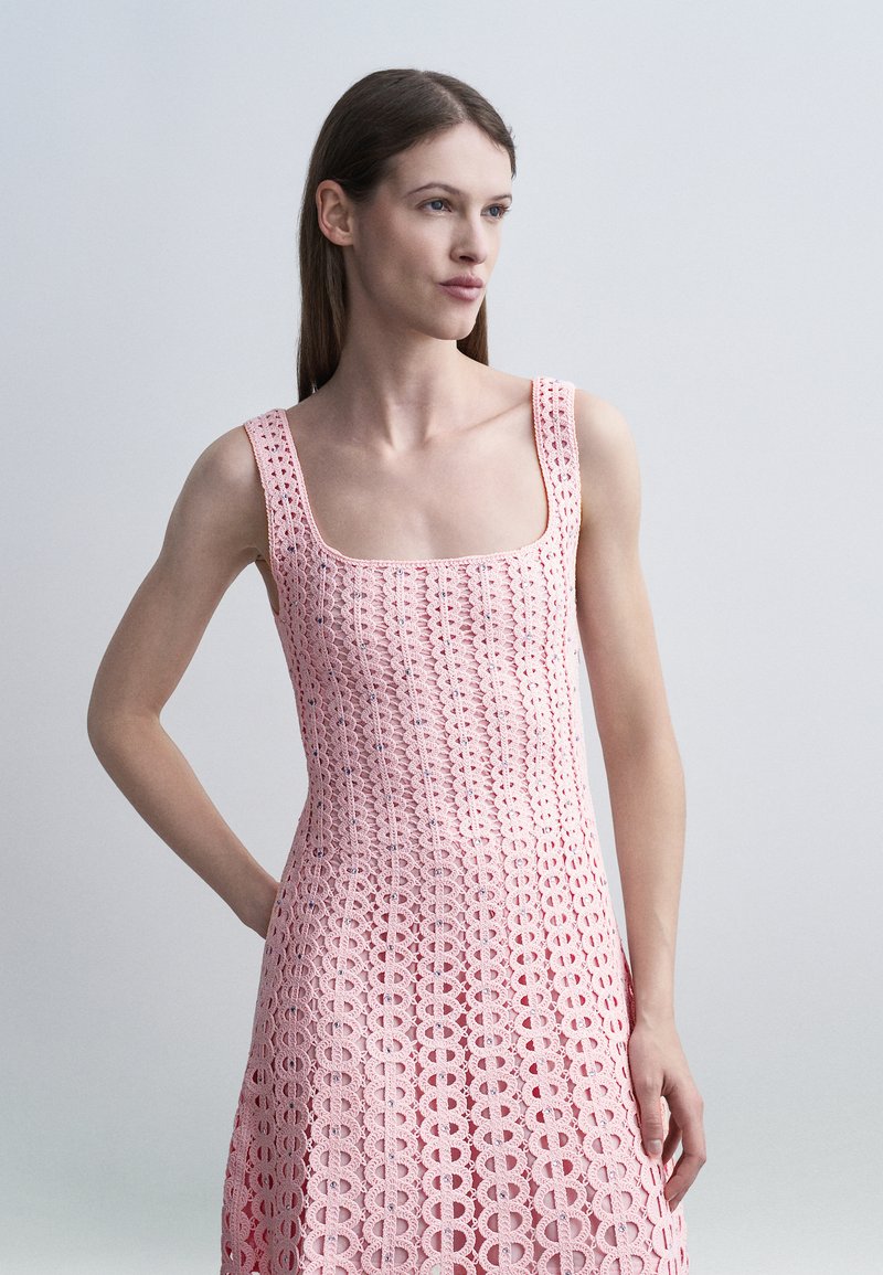 Femme portant une robe en crochet rose sans manches avec un col carré, debout avec une main sur la hanche devant un fond clair uni.